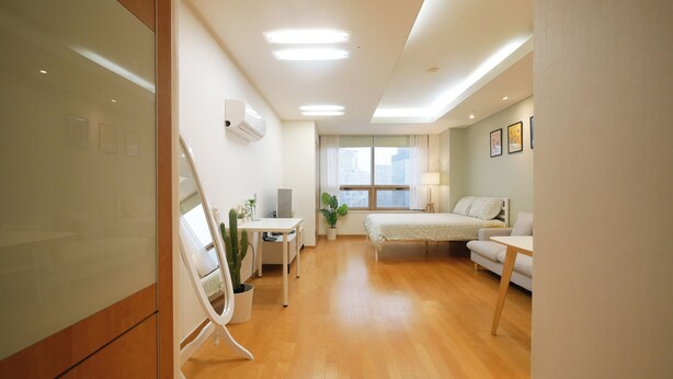 Une chambre d’hôtel moderne avec un plancher en bois, un lit recouvert d’une courtepointe à motifs floraux, un bureau avec un ordinateur, une chaise, une plante en pot et une fenêtre avec des rideaux.