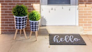 Een voordeur met een 'hello' mat, twee potplanten, een brievenbus en een jaartalplaatje.