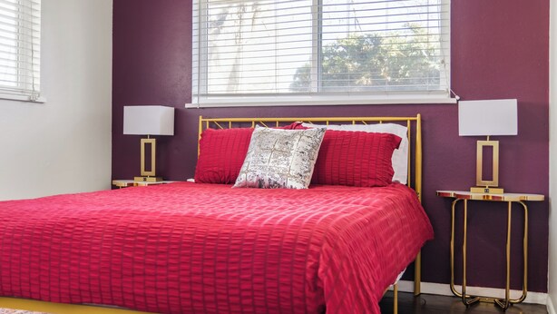 Een slaapkamer met een rode sprei, twee bedlampen, een gouden lijst rondom het hoofdeinde en een raam met jaloezieën.
