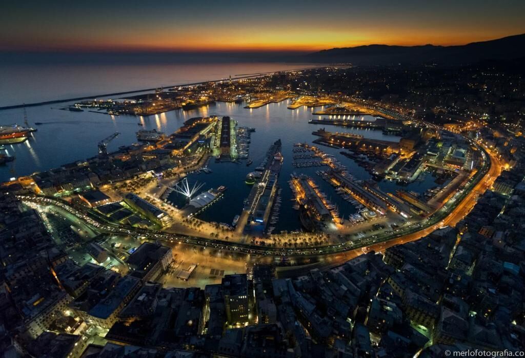 An aerial view of a coastal city at dusk with a marina, bridges, and illuminated buildings.