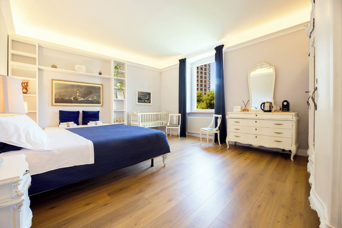 A bedroom with a bed, dresser, chair, and shelves with decorative items.