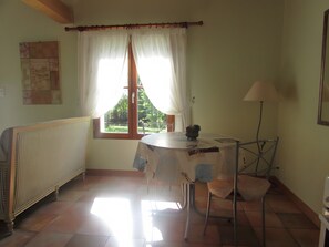 Una habitación con radiador, una mesa con un bol, una silla, una lámpara y una ventana con cortinas.
