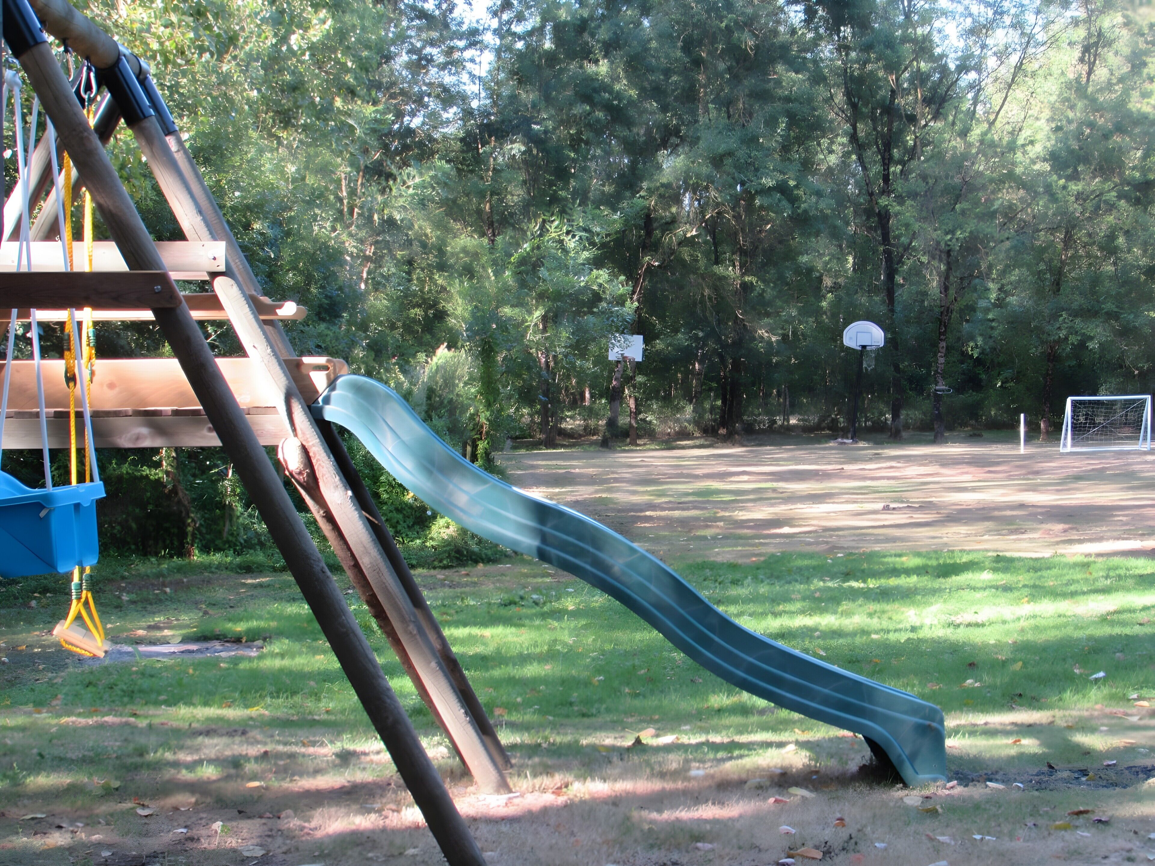 Un parque infantil con un tobogán, un aro de baloncesto y una portería de fútbol en una zona verde rodeada de árboles.