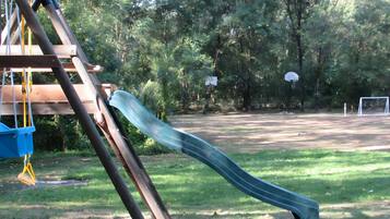 Un parque infantil con un tobogán, un aro de baloncesto y una portería de fútbol en una zona verde rodeada de árboles.