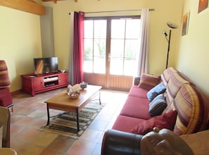 A living room with a sofa, a coffee table, a TV on a stand, and a window with curtains. - Gîte Sunflower Escape in Natura 2000: Marais Poitevin, Tranquility, Heated Pool, Wi-Fi (Nalliers)