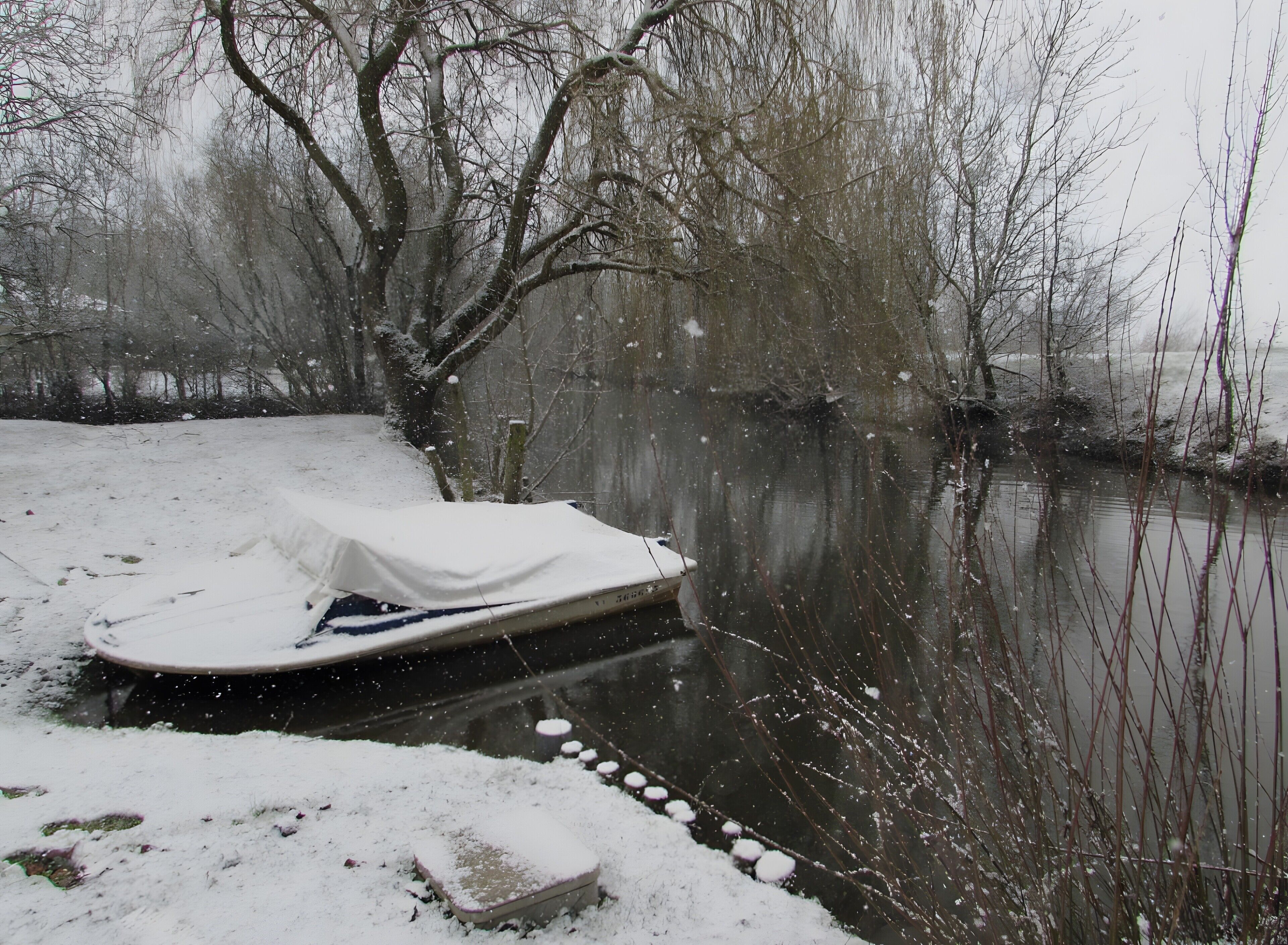 Un bateau recouvert d’une bâche blanche, amarré à un quai par une corde, avec de la neige au sol et des arbres en arrière-plan.