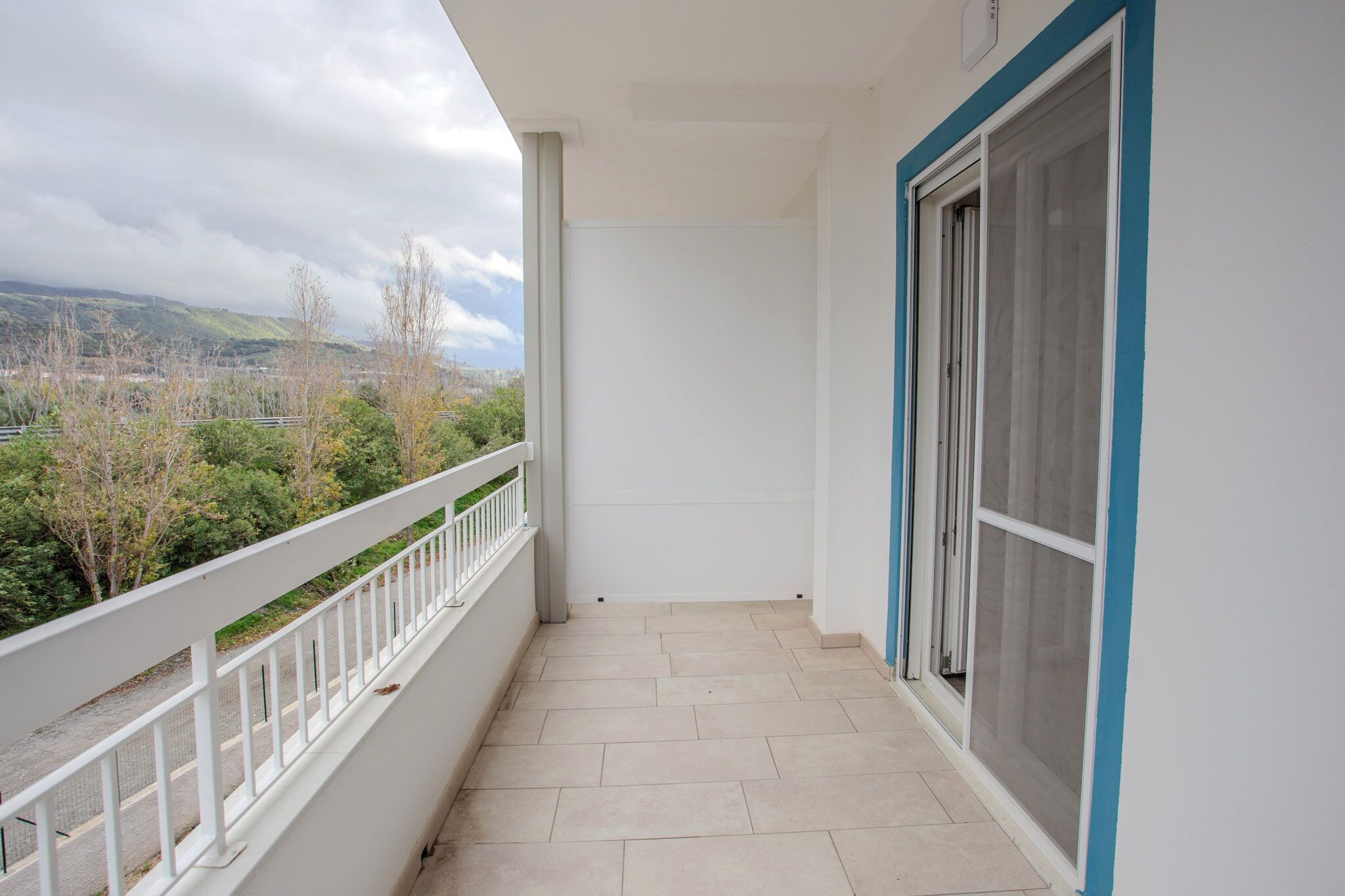 A balcony with a view of a cityscape and hills, white railings, and a sliding glass door.