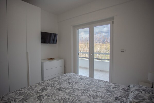 Un dormitorio con cama, cómoda, televisor montado en la pared y balcón con vista a la hierba alta.