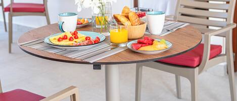 A breakfast table with plates of fruit, croissants, and orange juice. There are chairs around the table, and a bed with a blue pillow is visible in the background.
