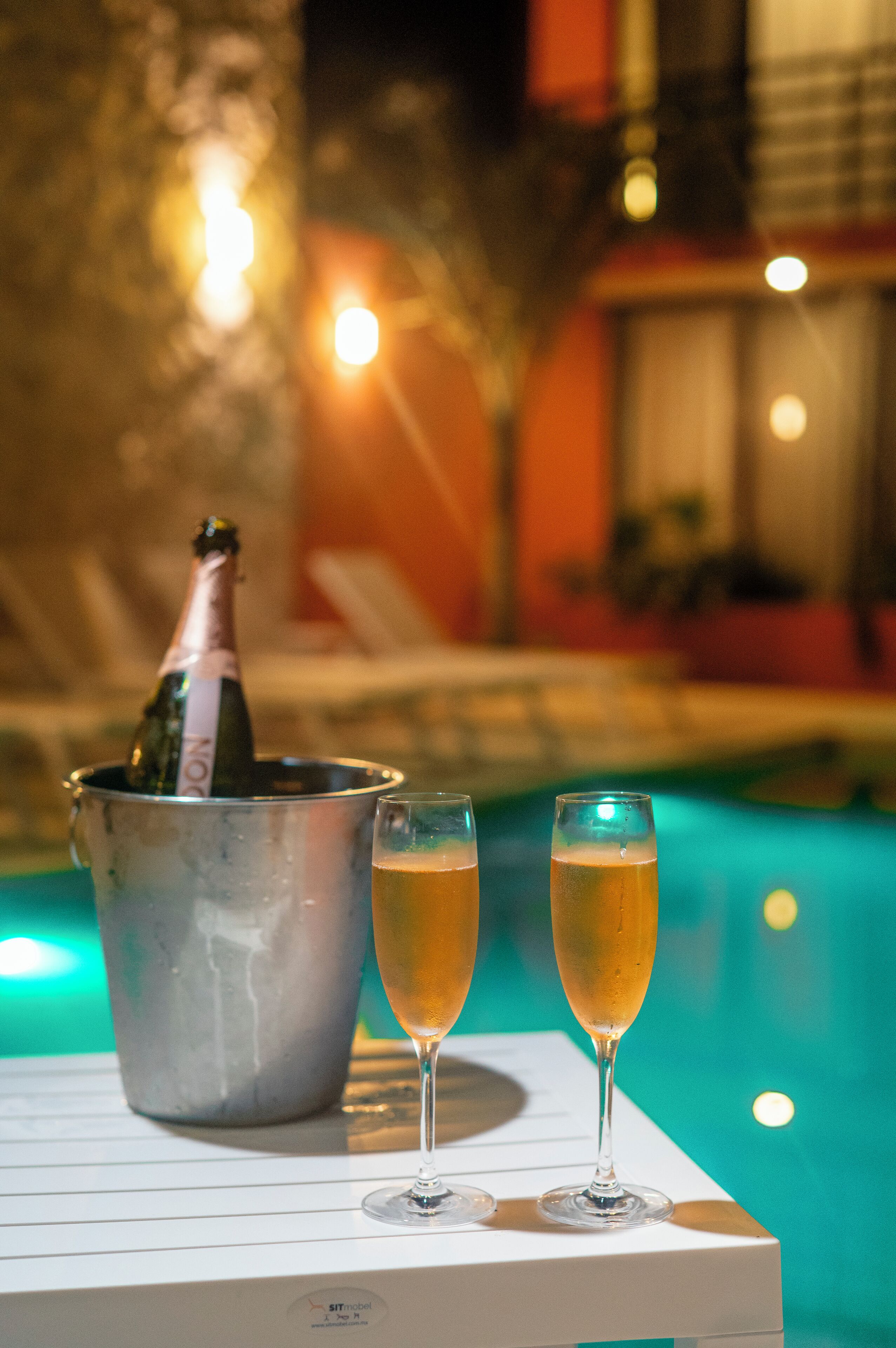 Escena junto a la piscina con una botella de champán en un cubo y dos copas de champán sobre una mesa blanca.