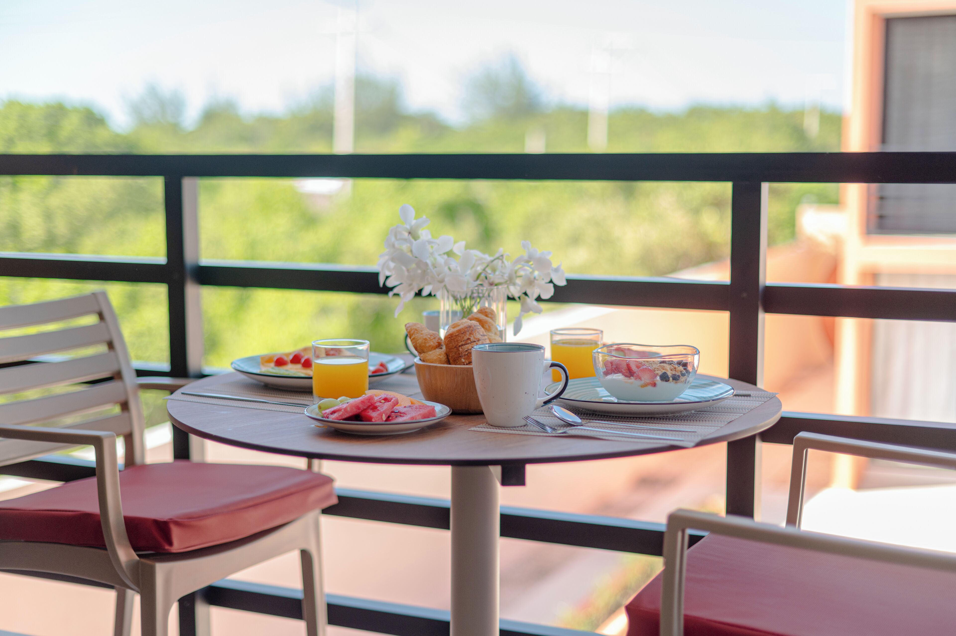 Une table de petit-déjeuner avec des assiettes remplies de nourriture, un vase de fleurs et une tasse de café, le tout placé sur un balcon donnant sur un paysage verdoyant.