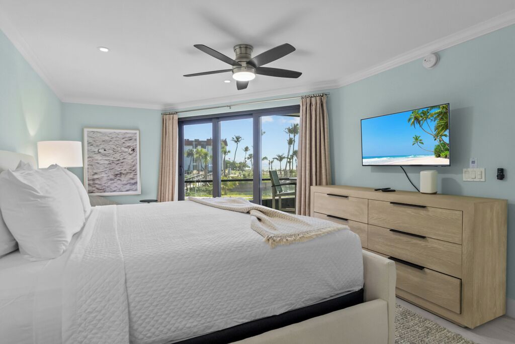 Un dormitorio con cama, ventilador de techo, televisión, ventana grande con cortinas y vista a palmeras.
