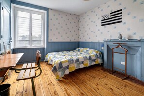 Quarto com cama, mesa e cadeira. Janela com persianas, parede com padrão decorativo e cômoda com espelho.