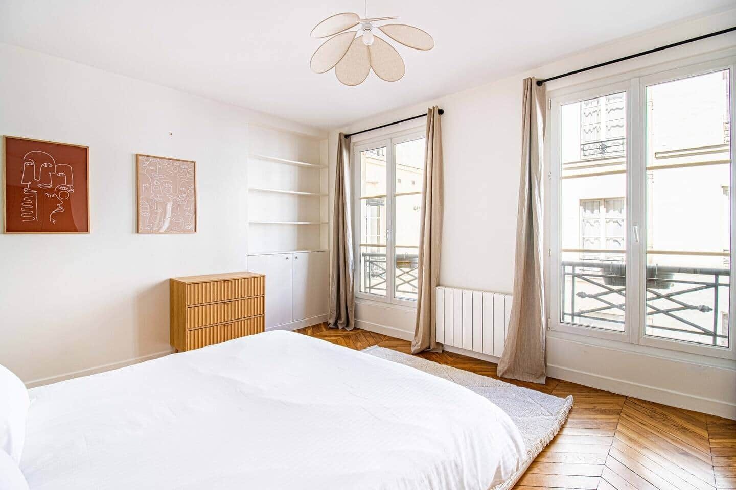 A bedroom with a bed, a wooden dresser, a ceiling fan, and large windows with curtains.