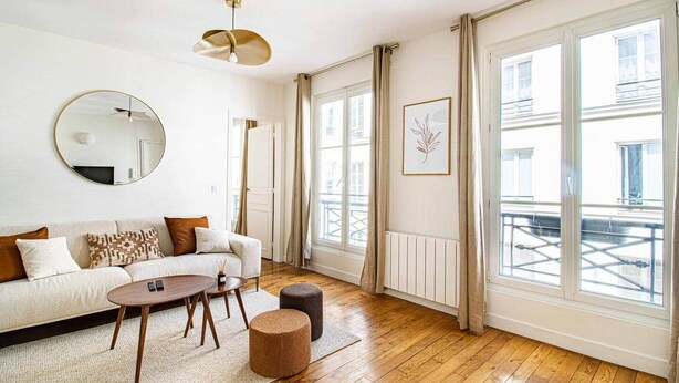 A modern living room with a sofa, coffee table, and ottoman, large windows with curtains, and a ceiling light fixture. - Sevres - Bright 2-bedroom near Bon Marché (Paris)
