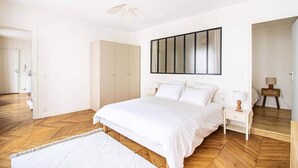 A bedroom with a large bed, wooden flooring, a wardrobe, a bedside table with a lamp, and a large window with curtains. - Sevres - Bright 2-bedroom near Bon Marché (Paris)