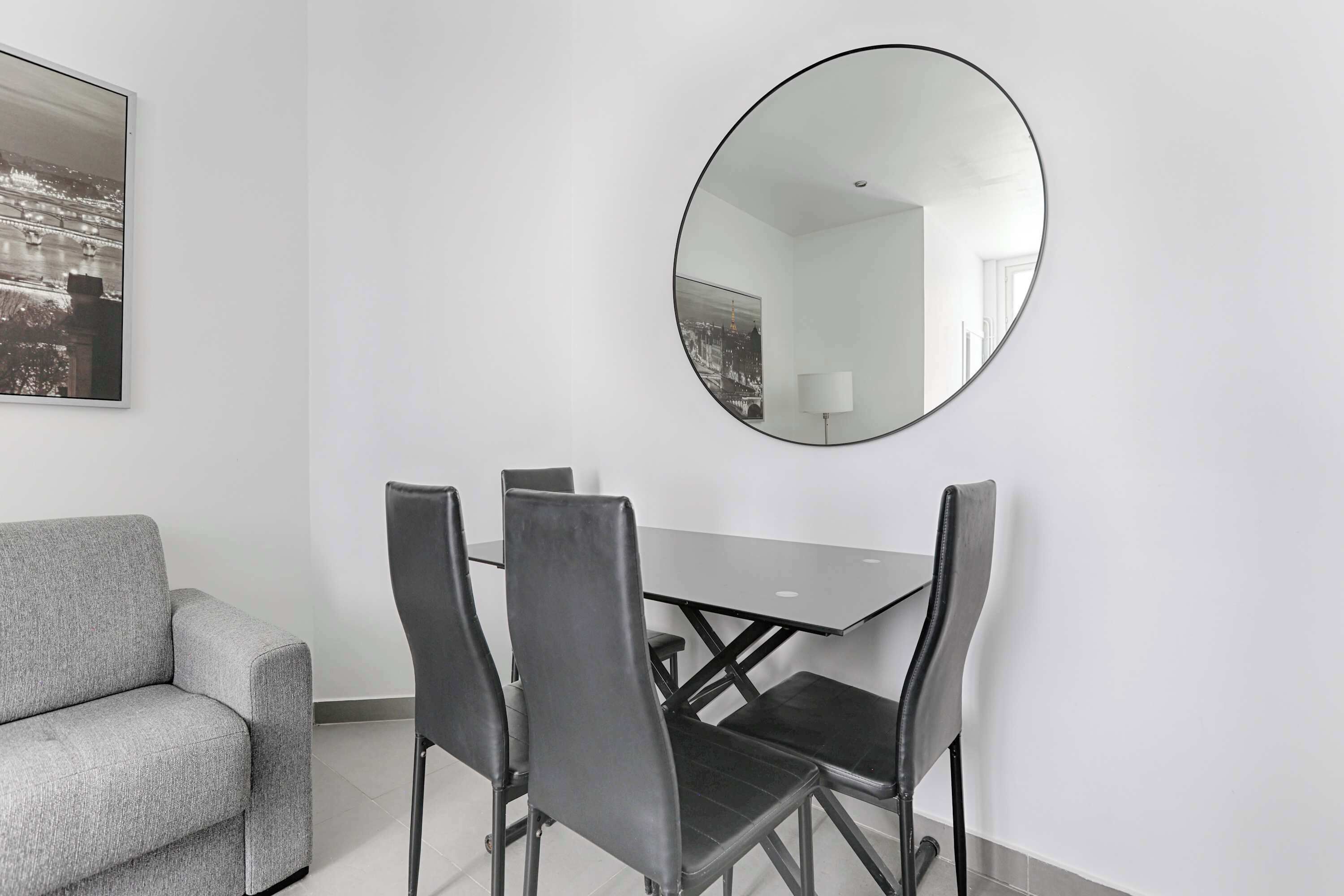 A modern room with a gray sofa, a dining table with four chairs, and a large round mirror on the wall.