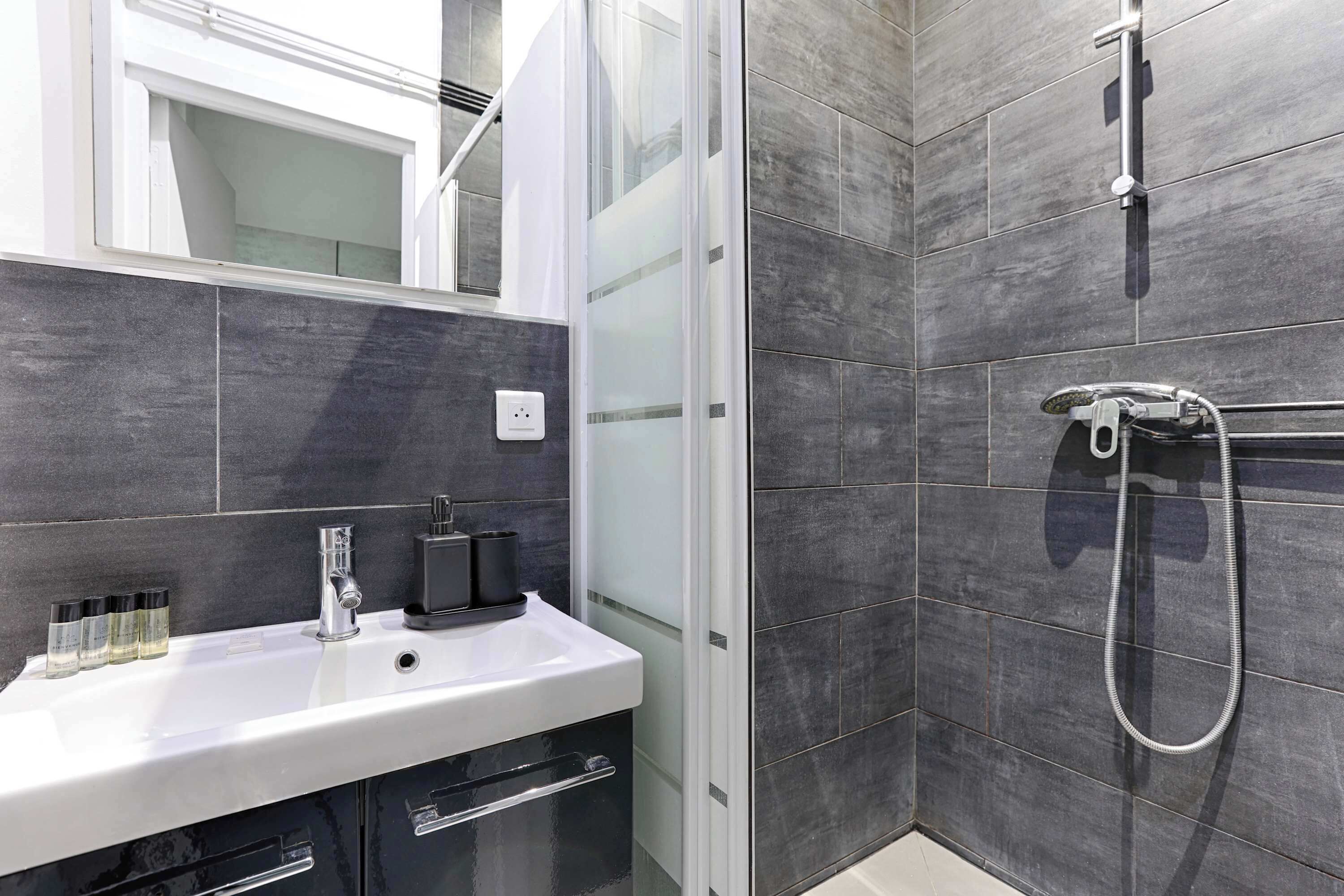A modern bathroom with a white sink, dark tiles, a glass shower enclosure, and a handheld showerhead.