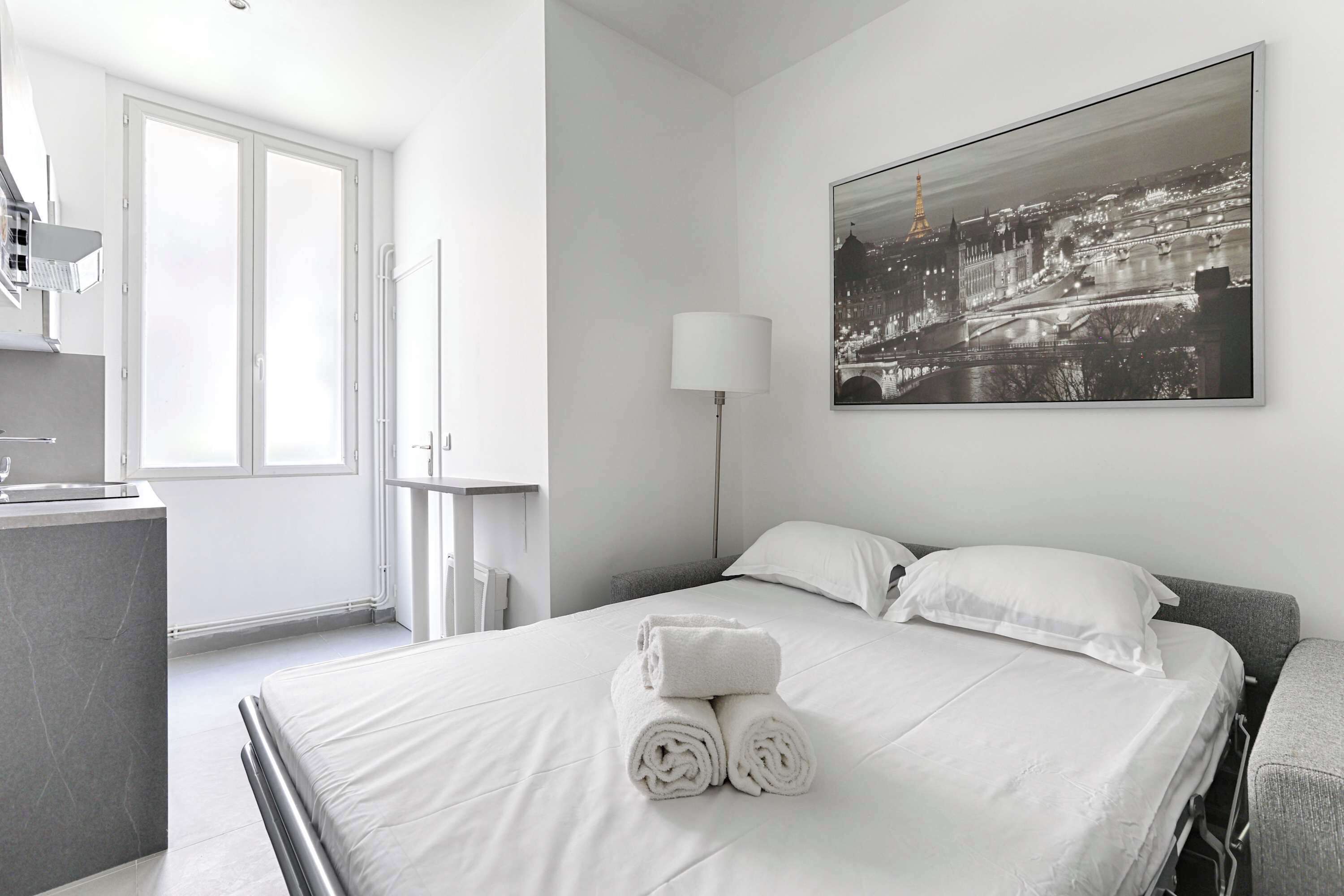 A hotel room with a bed, a large window, a desk with a chair, and a painting on the wall.