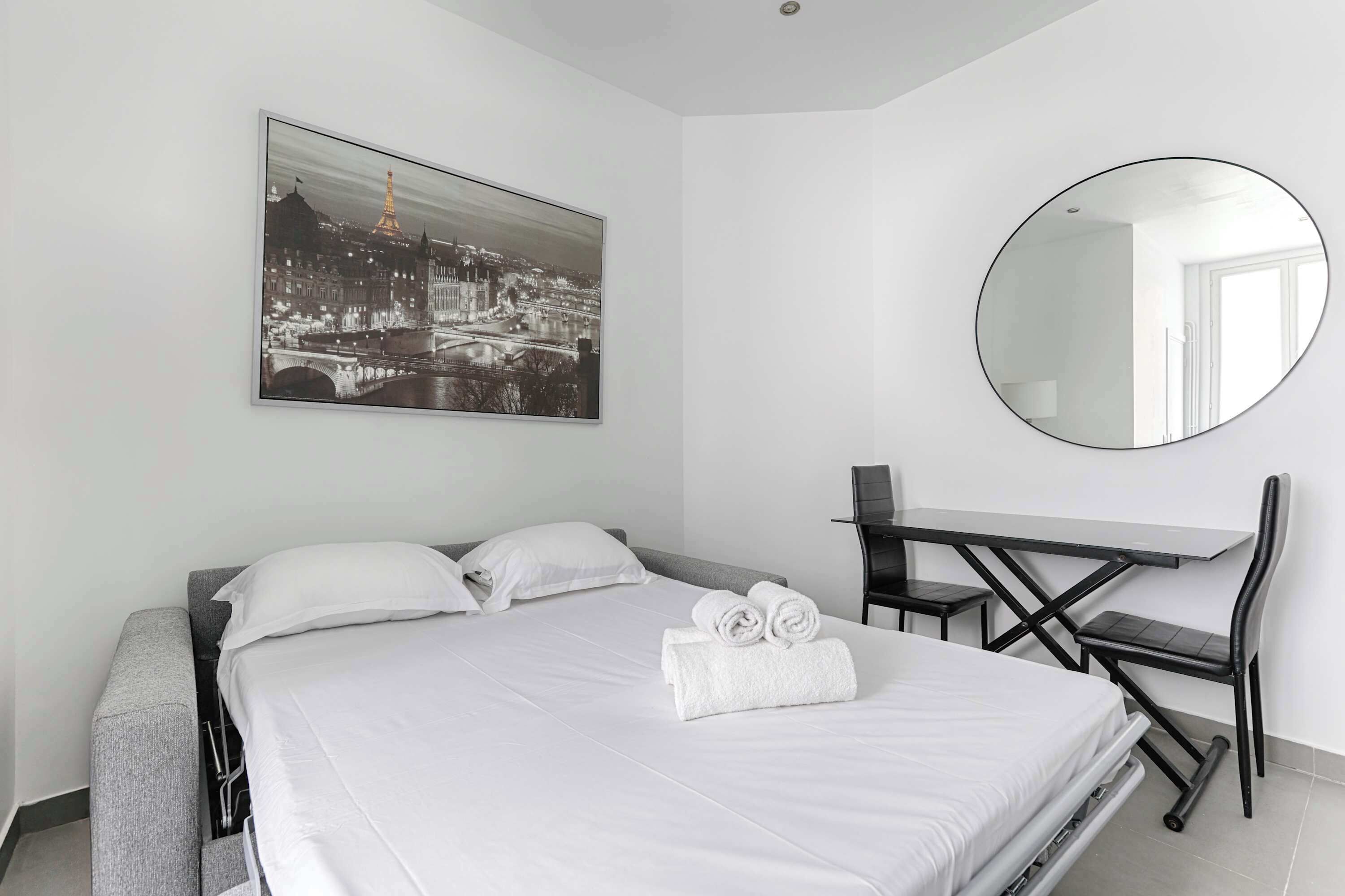 Ett hotellrum med en säng, ett skrivbord, en stol, en rund spegel och en tavla med en stadsbild på väggen.