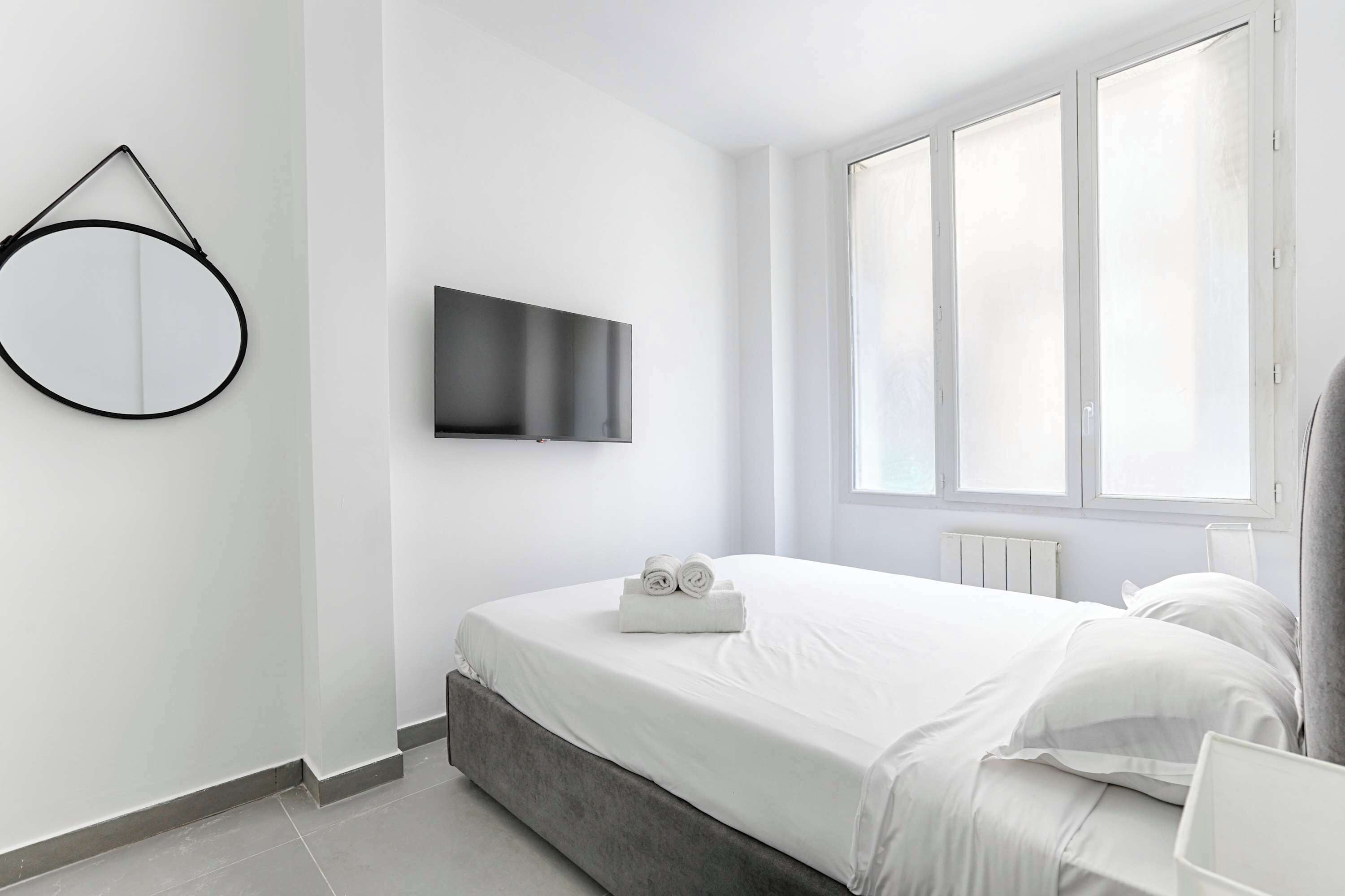 A modern bedroom with a bed, a TV mounted on the wall, and a window providing natural light.