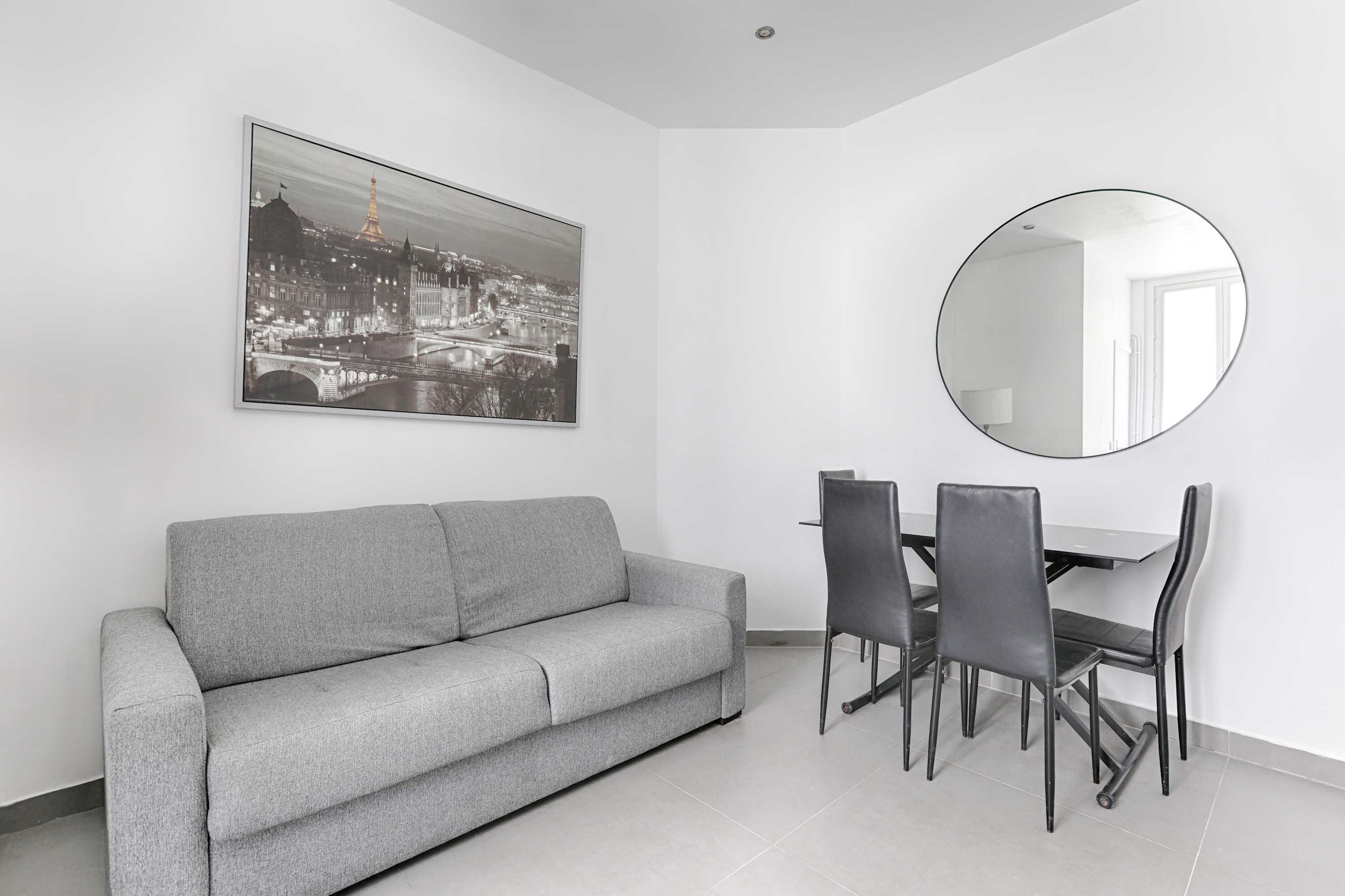 Un salon moderne avec un canapé gris, une table à manger avec quatre chaises, un miroir rond et un tableau représentant un paysage urbain en noir et blanc accroché au mur.