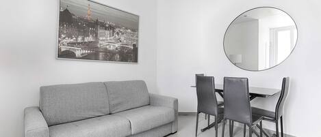 A modern living room with a grey sofa, a dining table with four chairs, a round mirror, and a black-and-white cityscape painting on the wall.
