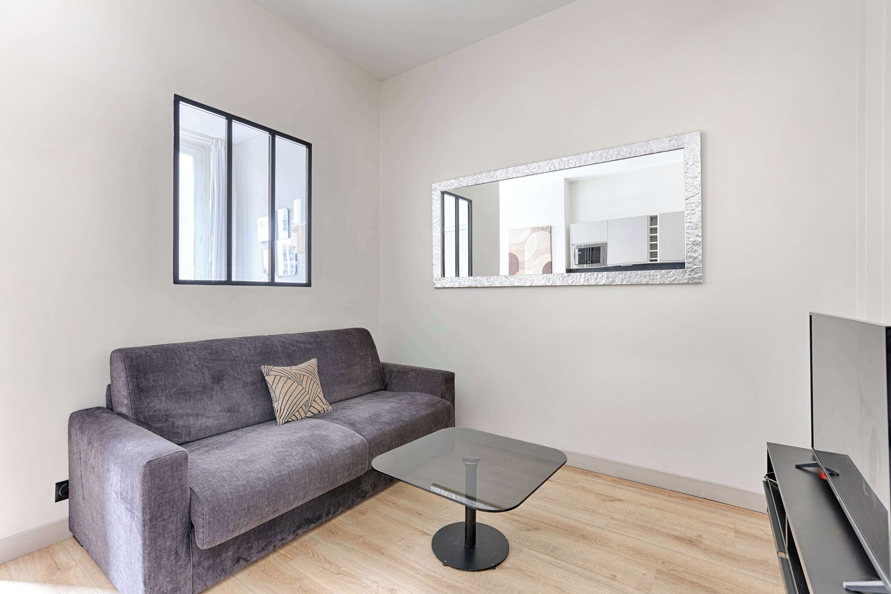 Un salon moderne avec un canapé gris, une table basse en verre, un grand miroir et un téléviseur à écran plat.
