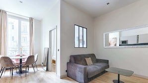 Un salón moderno con un sofá gris, una mesa de comedor con sillas, un espejo grande y vistas a la ciudad a través de una ventana.
