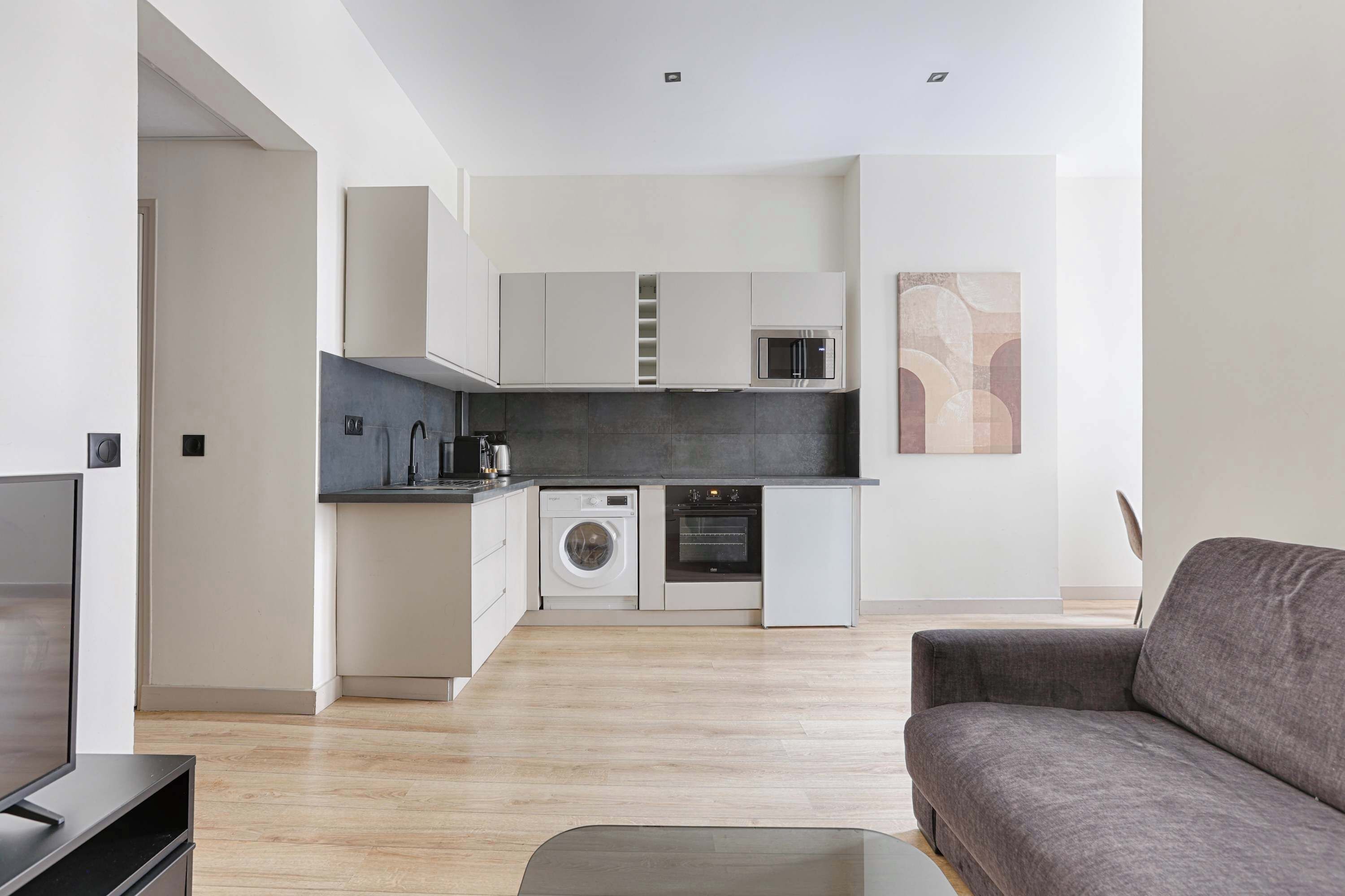 A modern kitchen with a microwave, oven, and washing machine, a grey sofa, and a flat-screen TV.