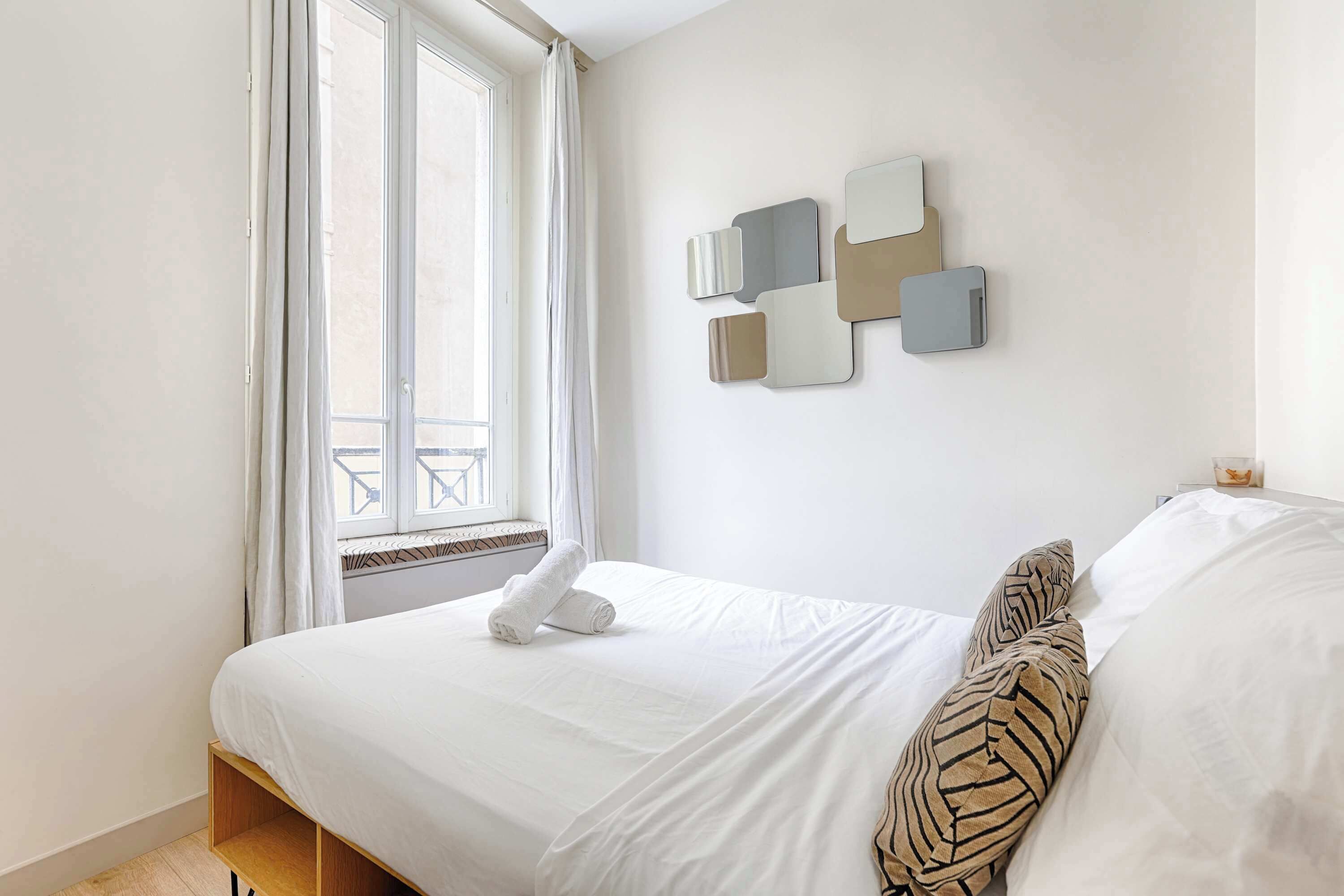 A neatly made bed with a white duvet and a rolled towel, a zebra-patterned pillow, and a wall-mounted abstract art piece.