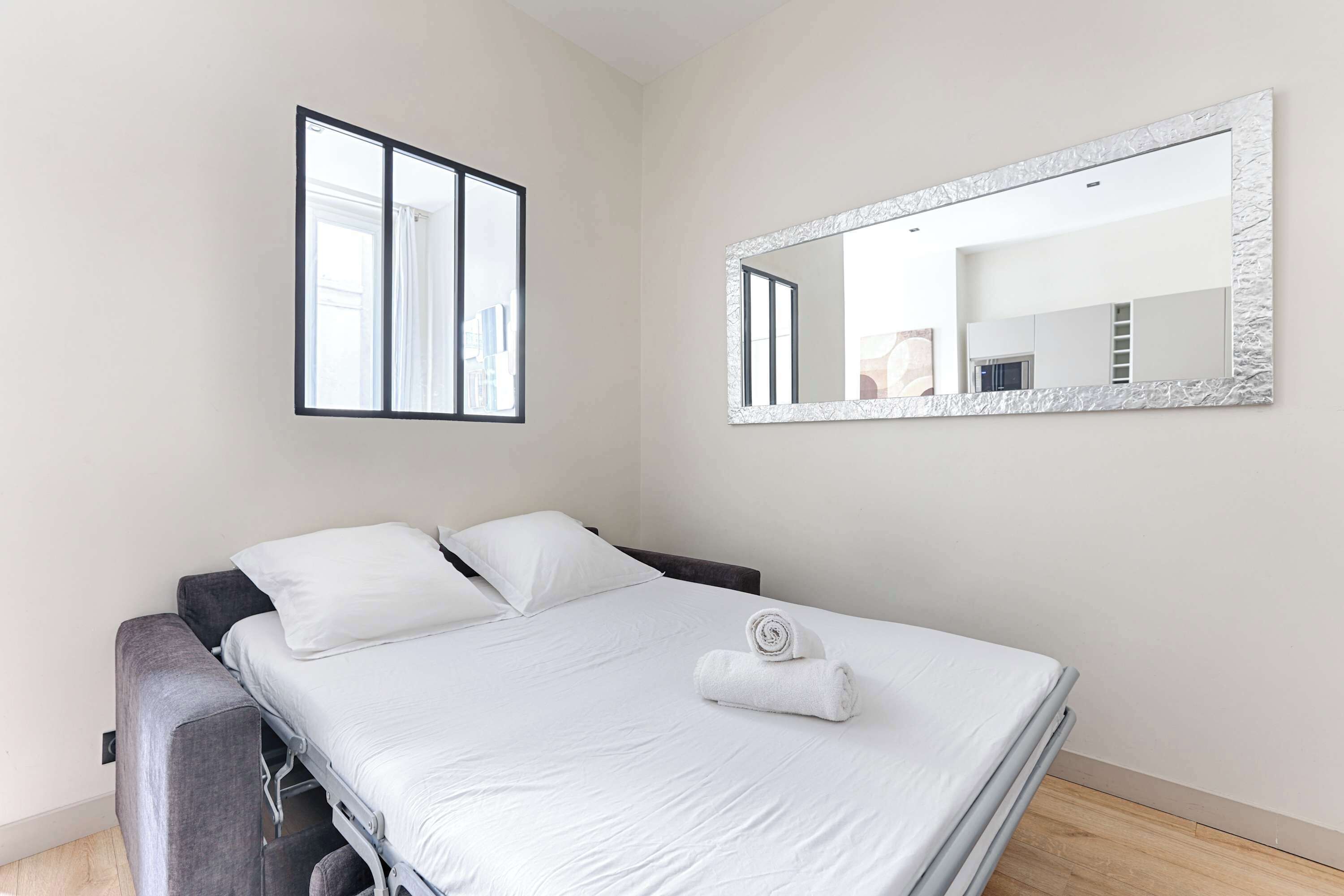 Un lit bien fait, avec du linge de lit blanc, un grand miroir doté d’un cadre texturé et une fenêtre avec des stores.