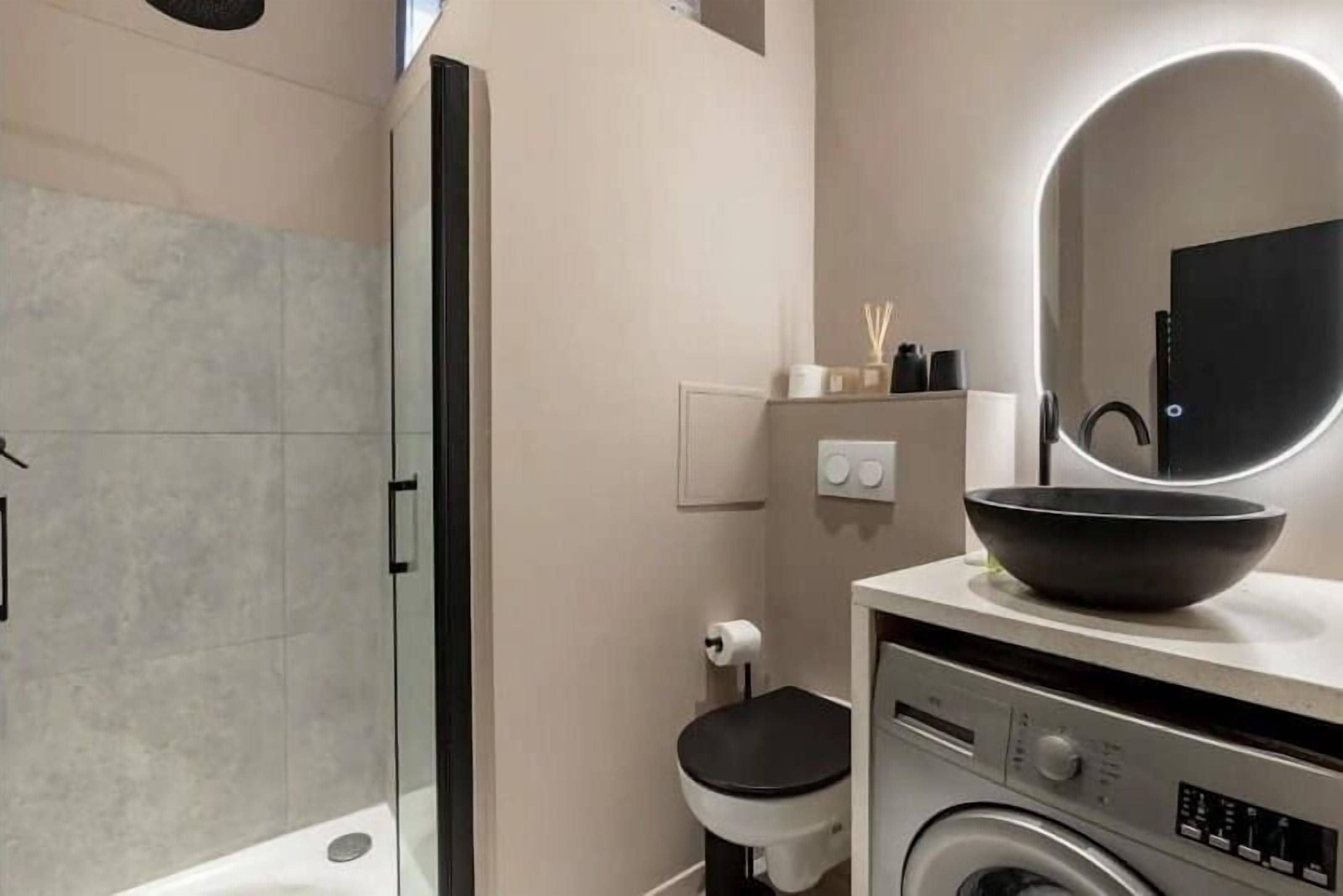 A modern bathroom with a glass shower enclosure, a toilet, a washing machine, and a sink with a bowl on the countertop.