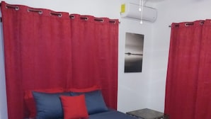 Un dormitorio con una cama, cortinas rojas, un ventilador de techo y un equipo de aire acondicionado, además de una mesita.
