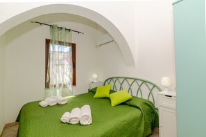 A bedroom with a green bedspread, two pillows, a nightstand with a lamp, and a window with a curtain. - San Teodoro Monti 22 – 20 minutes’ walk to the beach, private terrace & air conditioning (San Teodoro)