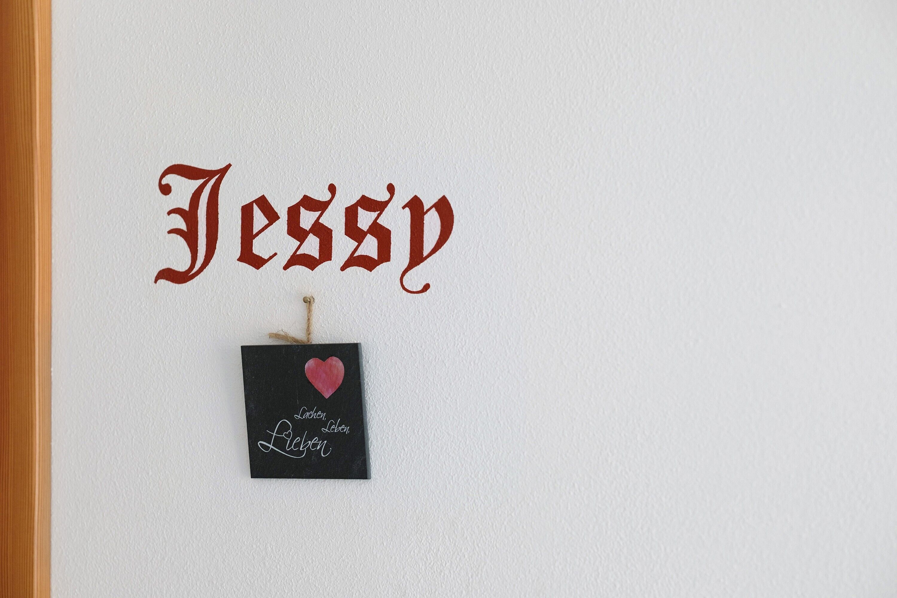 A wall with the name 'Jessy' in a decorative font, a small black sign with a heart and the words 'Lachen, Liebe, Leben' below it.