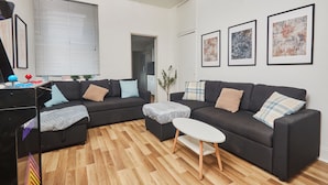 A modern living room with two black sofas, a small white table, and wooden flooring. There are framed artworks on the wall and a window with blinds. - Ladykirk Road (Newcastle-upon-Tyne)