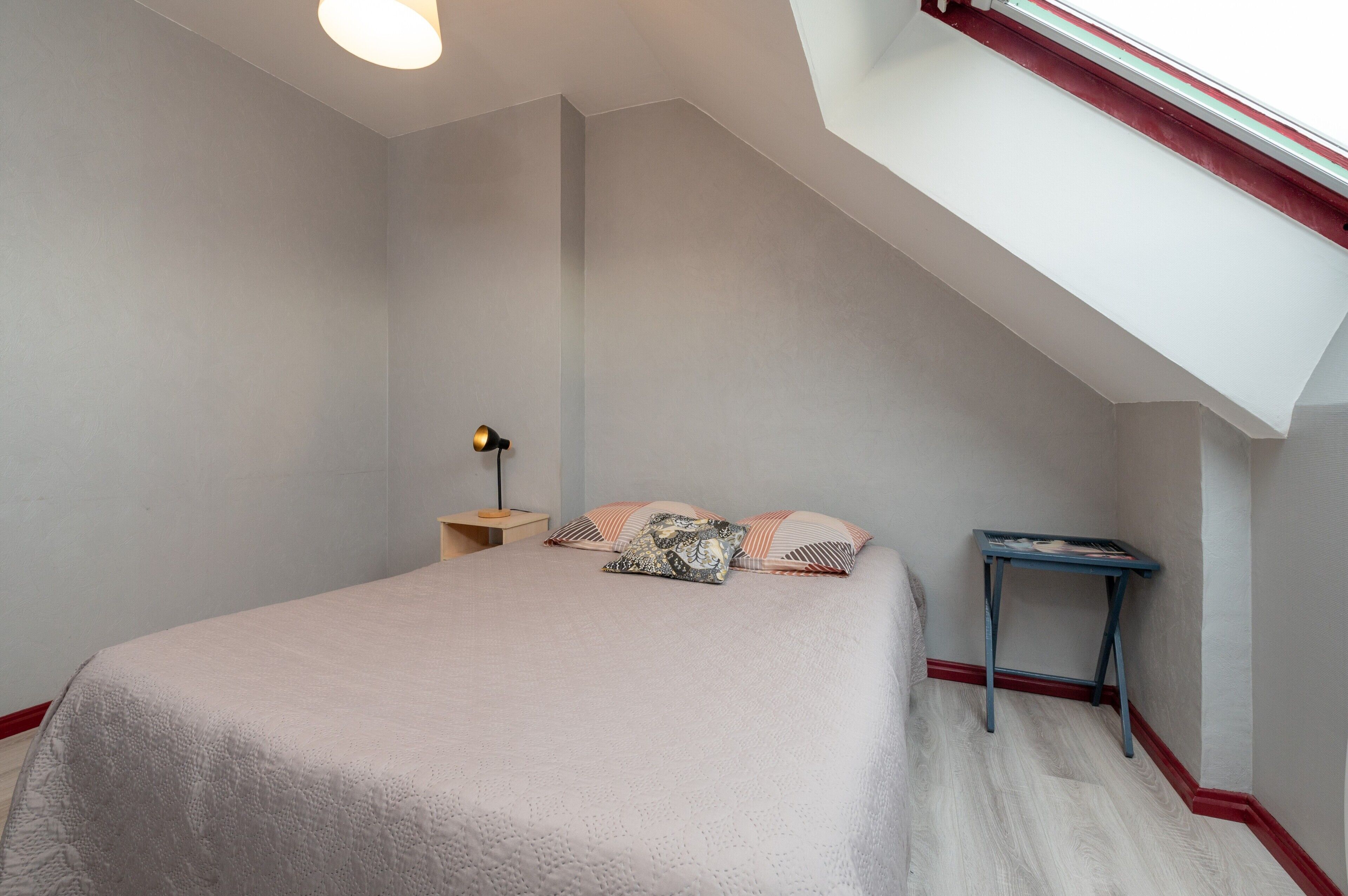 Une chambre à coucher avec un lit, des oreillers, une table de chevet, un bureau et une lucarne.