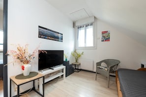 Une pièce dotée d’un téléviseur à écran plat, d’une petite table sur laquelle est posé un vase, d’une chaise en osier, d’un lit et d’une fenêtre avec des rideaux.