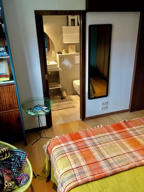 Une chambre à coucher avec un lit, un fauteuil vert, un panier à ouvrage et une salle de bain visible à travers une porte ouverte.