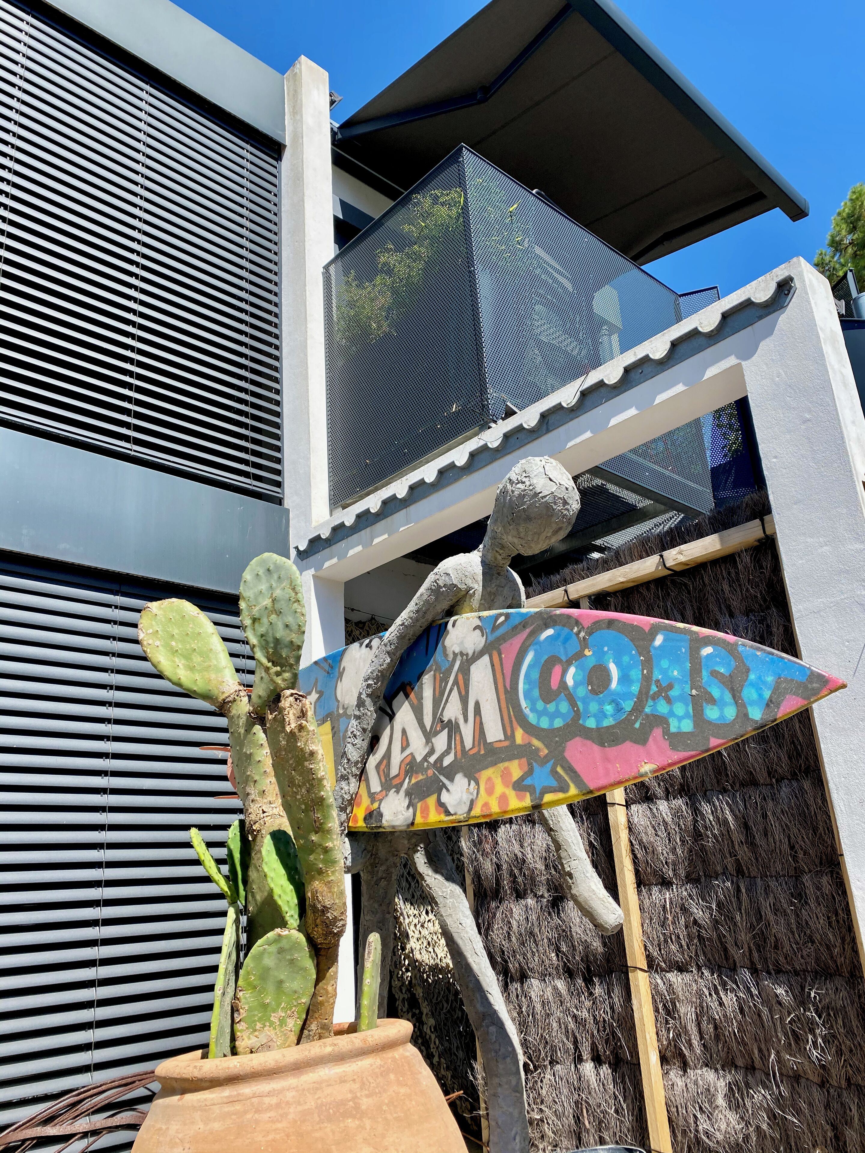 Ein Kaktus in einem Terrakotta-Topf, ein farbenfrohes Schild mit dem Schriftzug "Palm Coast" und eine moderne Gebäudefassade mit horizontalen Linien.