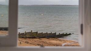 Blick durch ein Fenster auf einen Strand mit einem hölzernen Steg und ruhigem Meer.