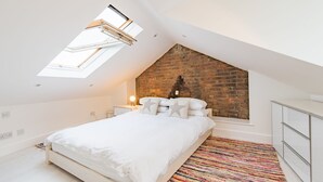 Ein Schlafzimmer mit geneigter Decke, einer Ziegelwand, einem Oberlicht, einem Bett mit weißer Bettwäsche, einem farbenfrohen Teppich und einer weißen Kommode.