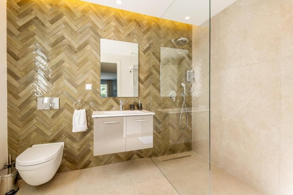 Et moderne badeværelse med en væg i fiskebensmønstret træ, et hvidt toilet, et vaskeskab med spejl og et bruseområde med glasvægge.