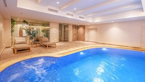 A spacious indoor swimming pool area with lounge chairs and a glass wall featuring plants.