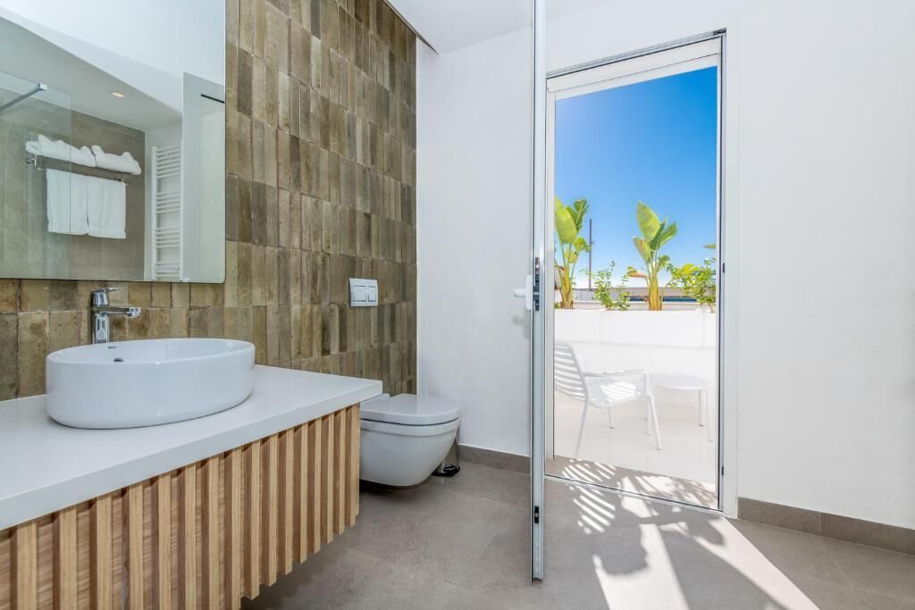 A modern bathroom with a large sink, toilet, and a glass door leading to an outdoor area with chairs and plants.