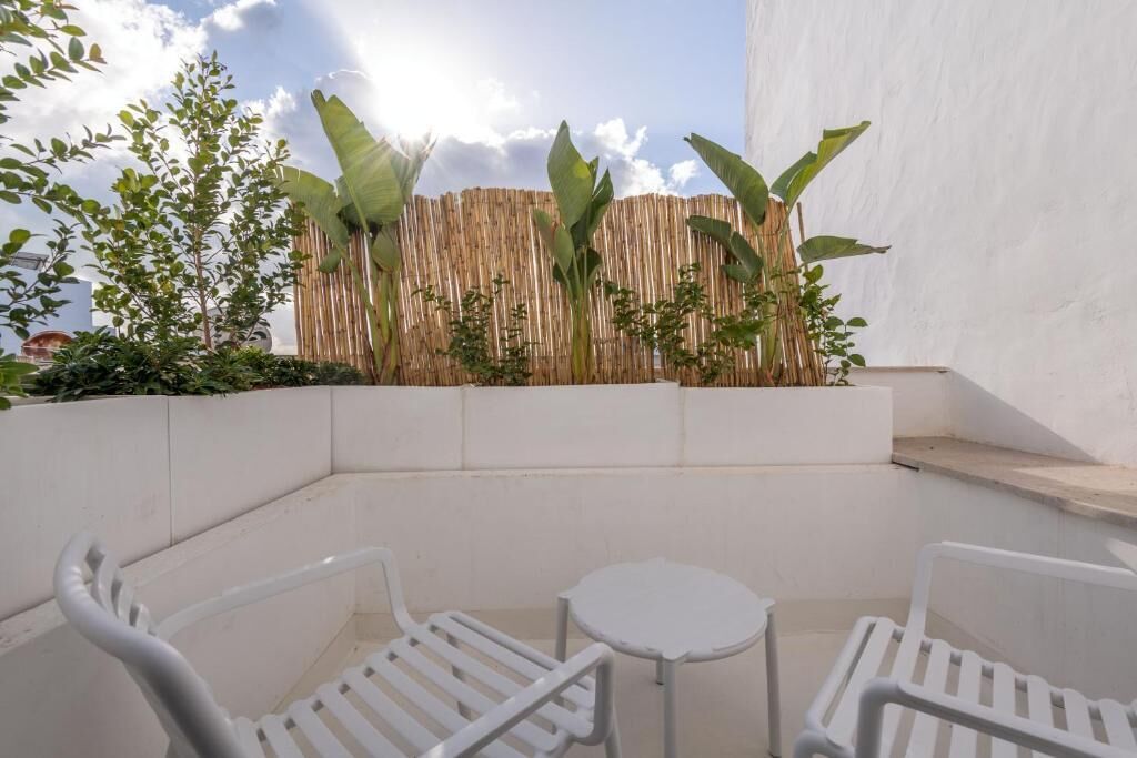 Un petit espace extérieur aménagé avec des chaises en plastique blanc et une table, entouré de plantes et d’une clôture en bambou.