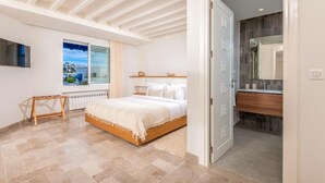 A bedroom with a bed, a wooden bench, a window, and a bathroom with a mirror and sink.