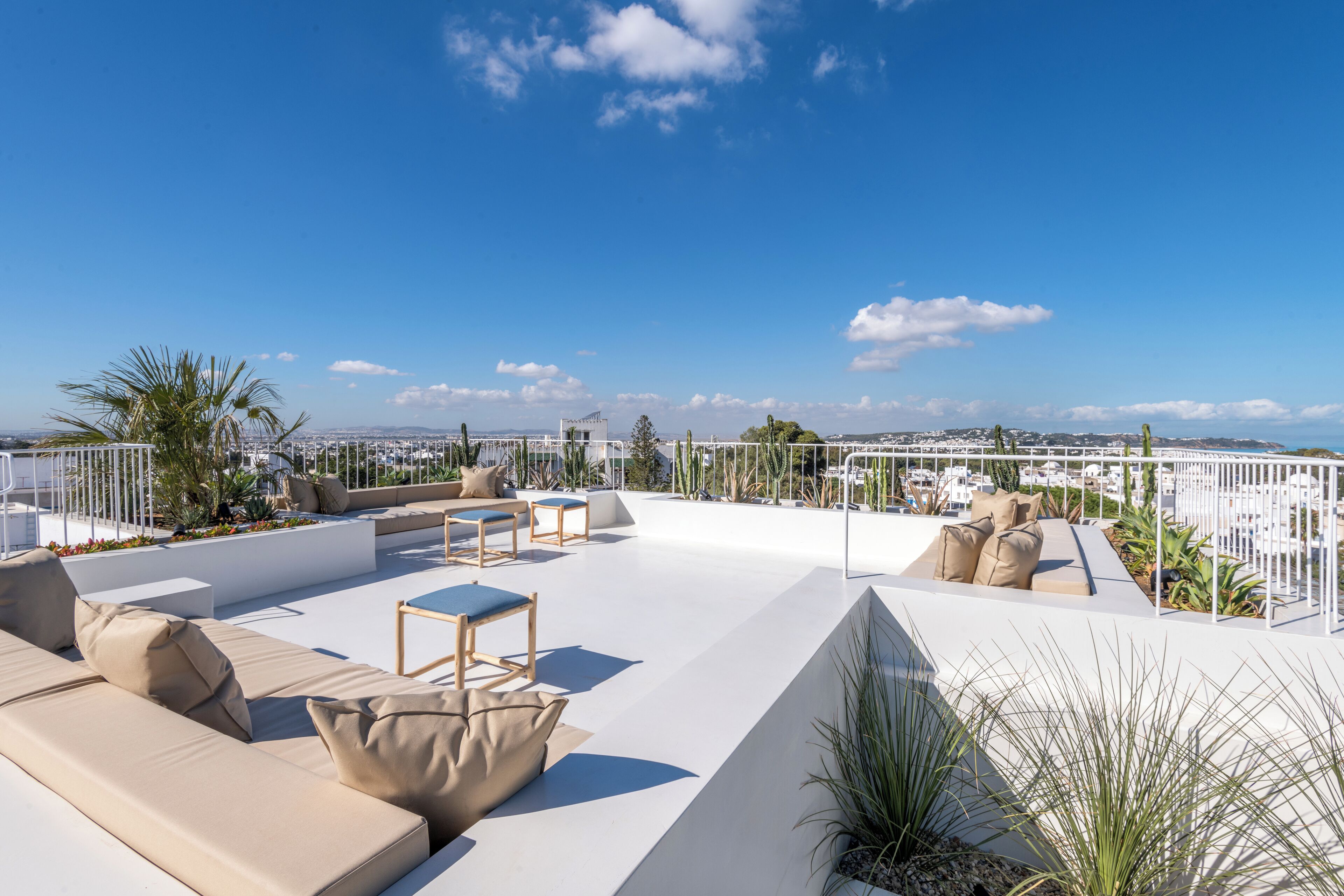 Una terraza en la azotea con zona de estar exterior blanca, que incluye sofás y sillones, rodeada de plantas y una vista despejada del paisaje urbano.