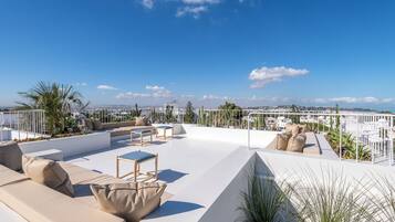 Una terraza en la azotea con zona de estar exterior blanca, que incluye sofás y sillones, rodeada de plantas y una vista despejada del paisaje urbano.